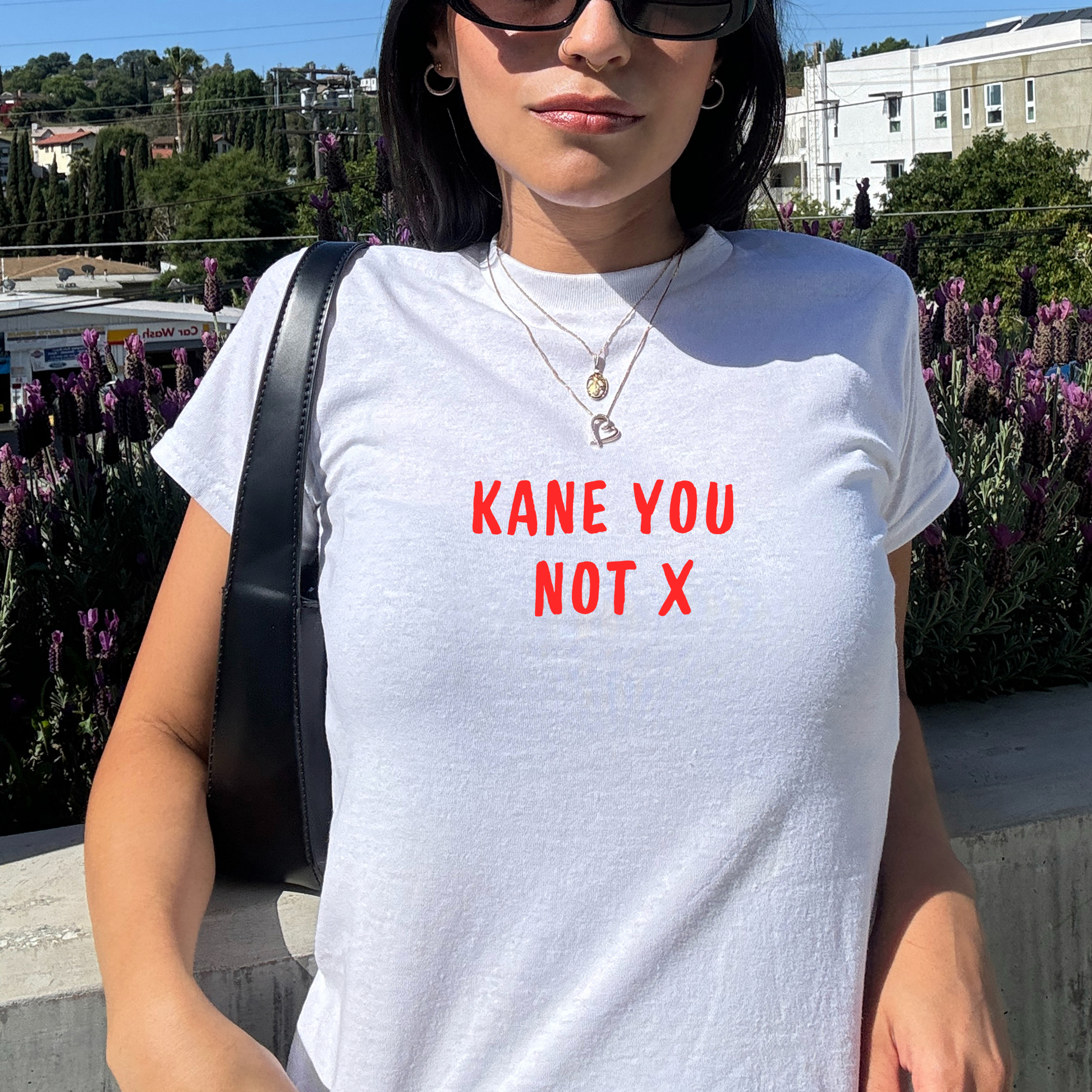 White fitted baby tee with red “Kane You Not X” slogan, styled casually with a shoulder bag, perfect for England football fans and match day outfits