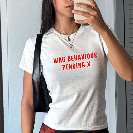 White fitted baby tee with red “WAG Behaviour Pending X” slogan, styled casually with a shoulder bag, perfect for football fans and match day outfits