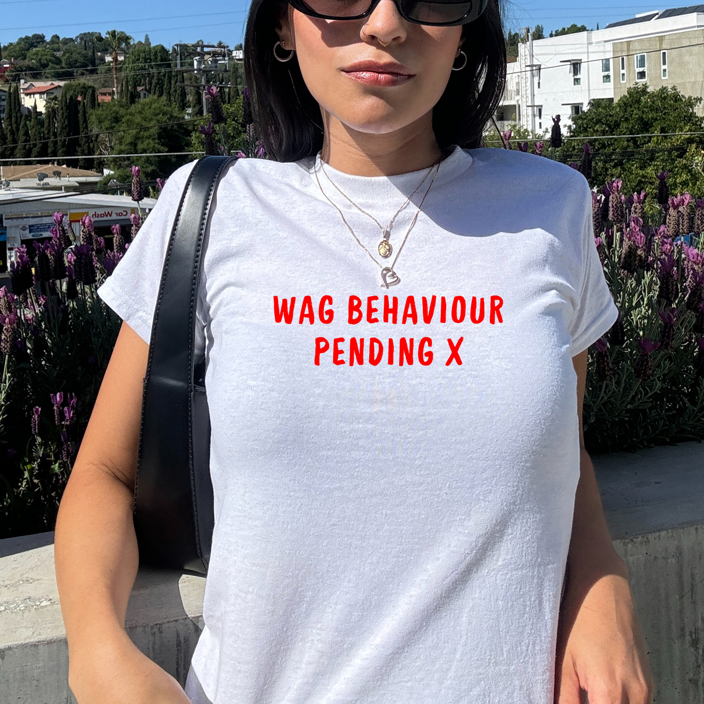White fitted baby tee with red “WAG Behaviour Pending X” slogan, styled casually with a shoulder bag, perfect for football fans and match day outfits