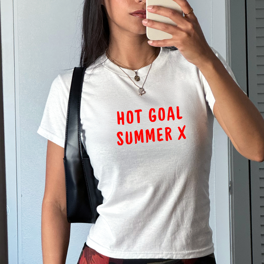White fitted baby tee with bold red “Hot Goal Summer X” slogan, styled casually with a shoulder bag, ideal for summer football outfits and match days